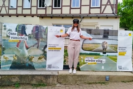 Erxleben © MID Frau mit Fahrradhelm zeigt auf aufjepasst-Banner für Rücksicht auf Kinder und Abstand im Straßenverkehr.
